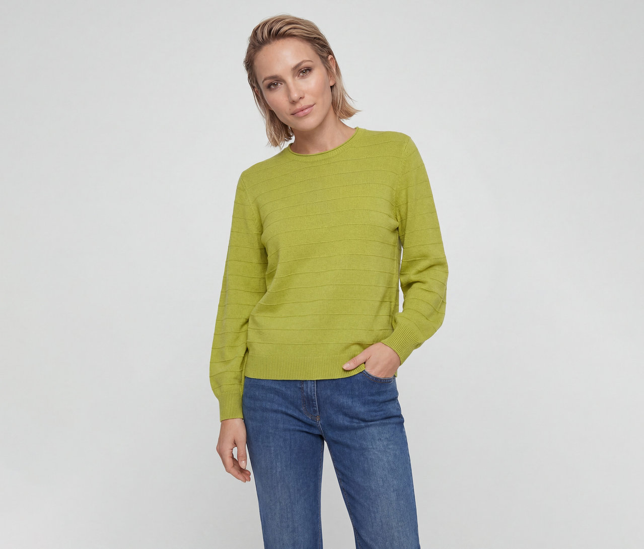 Frau mit kurzen blonden Haaren trägt einen limettengrünen Pullover mit horizontalen Strukturstreifen und blaue Jeans, blickt in die Kamera, eine Hand in der Tasche.