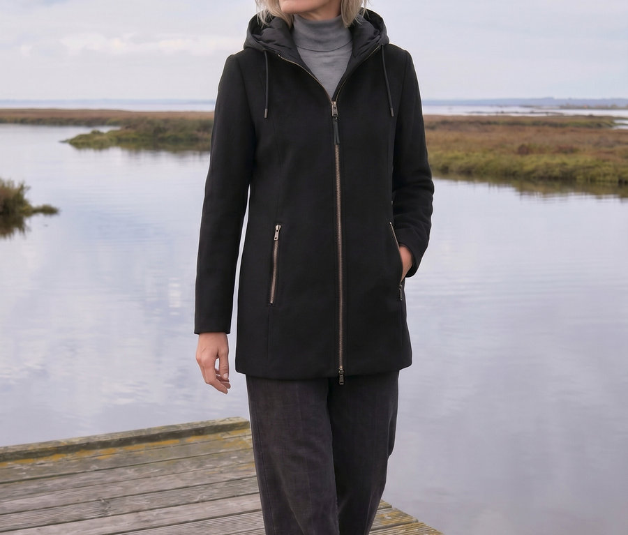 Eine Frau mit hellem Haar geht auf einem Holzsteg am Wasser entlang. Sie trägt eine schwarze Kapuzenjacke, einen grauen Rollkragenpullover und dunkle Hosen und blickt nach rechts. Im Hintergrund sind Marschland und ein bewölkter Himmel zu sehen.