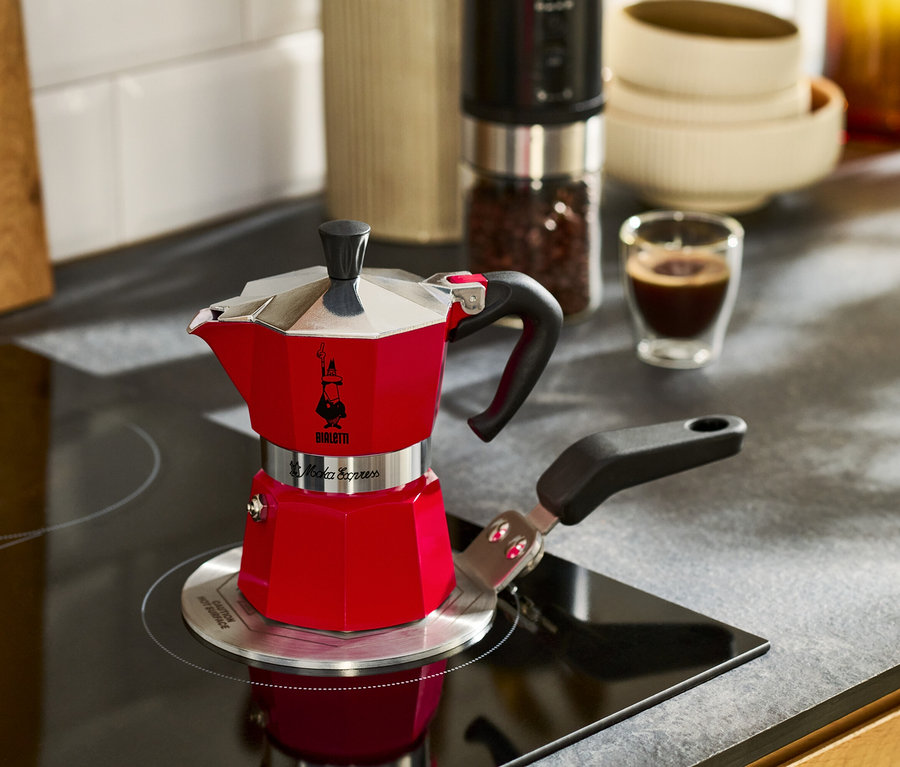 Ein roter Espressokocher steht auf einem Kochfeld neben einer Tasse Espresso.