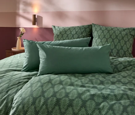 Ein Bett mit grüner Bettwäsche, die ein Farnblattmuster hat, und vier Kissen. Im Hintergrund ist eine zweifarbige Wand in Lila- und Rosatönen zu sehen, daneben ein Nachttisch mit Vase und Blume sowie eine Wandleuchte.