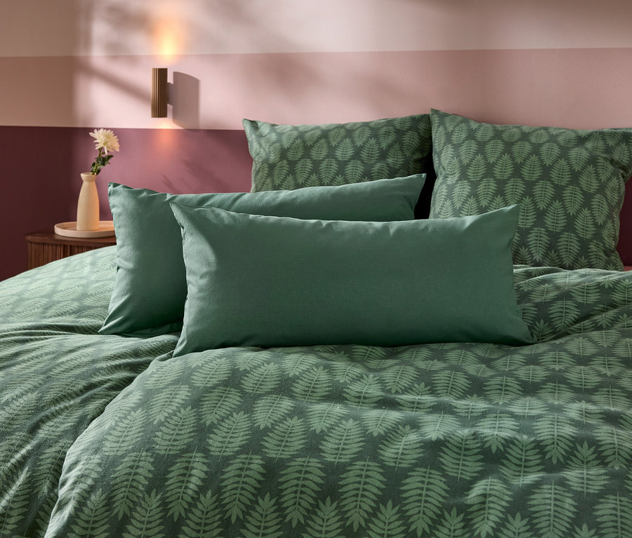 Ein Bett mit grüner Bettwäsche, die ein Farnblattmuster hat, und vier Kissen. Im Hintergrund ist eine zweifarbige Wand in Lila- und Rosatönen zu sehen, daneben ein Nachttisch mit Vase und Blume sowie eine Wandleuchte.