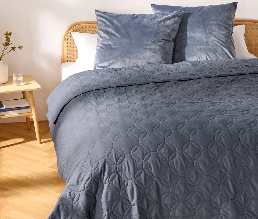 Ein Bett mit hellem Rattan-Kopfteil, bedeckt mit einer dunkelblauen gesteppten Tagesdecke mit kreisförmigem Muster. Zwei hellblaue Samtkissen liegen am Kopfteil. Links steht ein heller Nachttisch mit einer weißen Vase mit Pflanze und einem Glas Wasser. Der Raum hat helles Parkett.