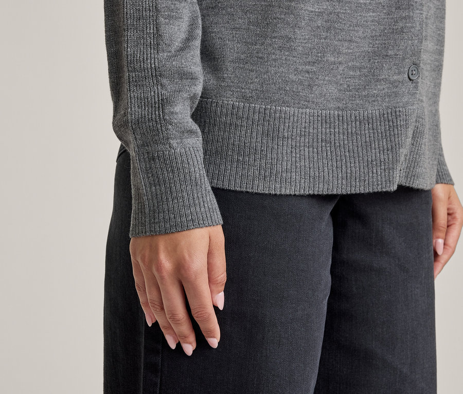Nahaufnahme einer Person in einem grauen Langarmshirt mit gerippten Bündchen und Saum sowie einer dunklen Hose. Ein kleiner dunkler Knopf ist seitlich am Oberteil sichtbar. Die Hände der Person haben hellrosa Nagellack.