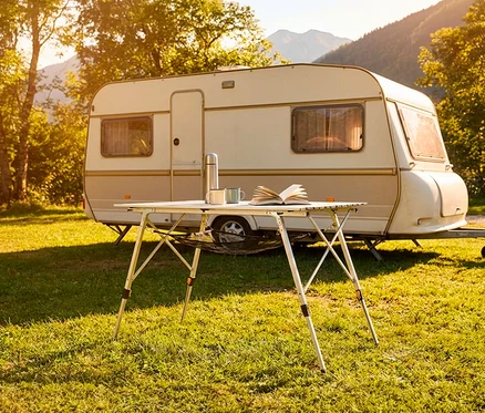 Ein Faltbarer Campingtisch steht vor einem Wohnwagen auf einer sonnigen Wiese mit Bergen im Hintergrund. Auf dem Tisch liegen ein Buch, eine Thermoskanne und zwei Tassen.