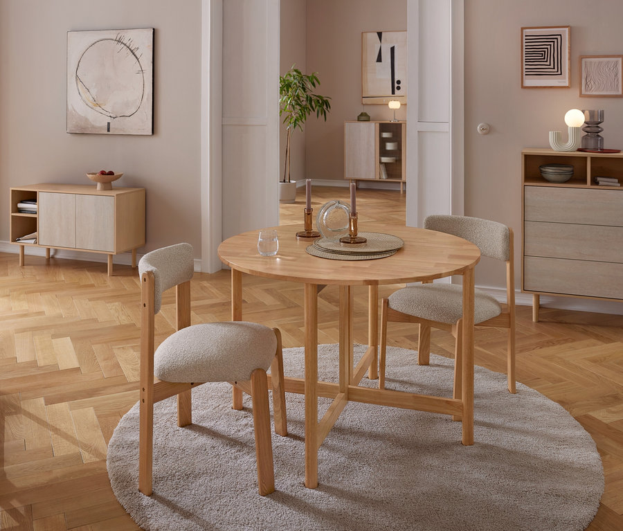 Ein Essbereich mit einem runden Holztisch und drei hell gepolsterten Stühlen auf einem grauen Teppich. Der Tisch ist mit Kerzenständern und einem Glas gedeckt. Im Hintergrund stehen ein Holz-Sideboard und eine Kommode an neutralen Wänden mit abstrakter Kunst, alles auf Parkettboden.
