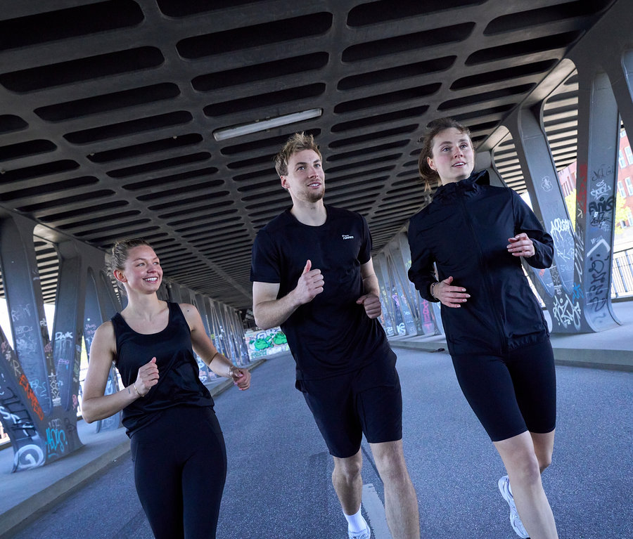 Drei Personen laufen unter einer Brücke: Eine Frau in schwarzem Sport-Top und schwarzen Sportleggings »ActivePower«, ein Mann in schwarzem Sportshirt und schwarzen Sportshorts, und eine Frau in schwarzer Windjacke »packable« und schwarzen Sportradler »ActivePower«.