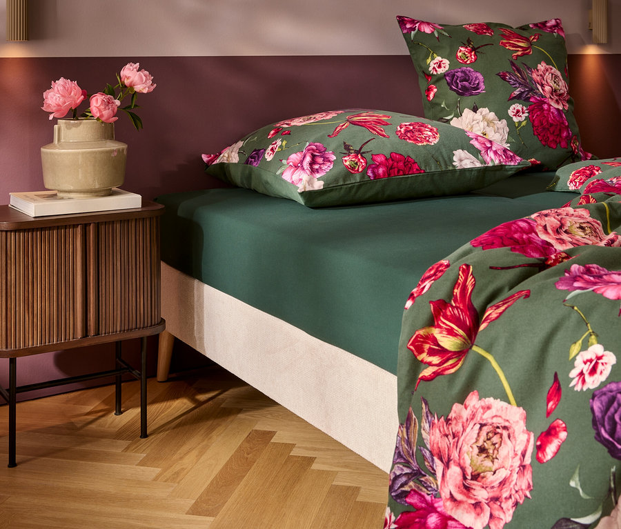 Ein Bett mit dunkelgrüner Bettwäsche und lebhaftem Blumenmuster in Pink, Fuchsia und Lila. Links steht ein Nachttisch aus Holz mit einer Vase voller pinker Pfingstrosen und einem Buch. Die Wand ist zweifarbig in Pflaume und Hellrosa, über einem Fischgrätparkett.