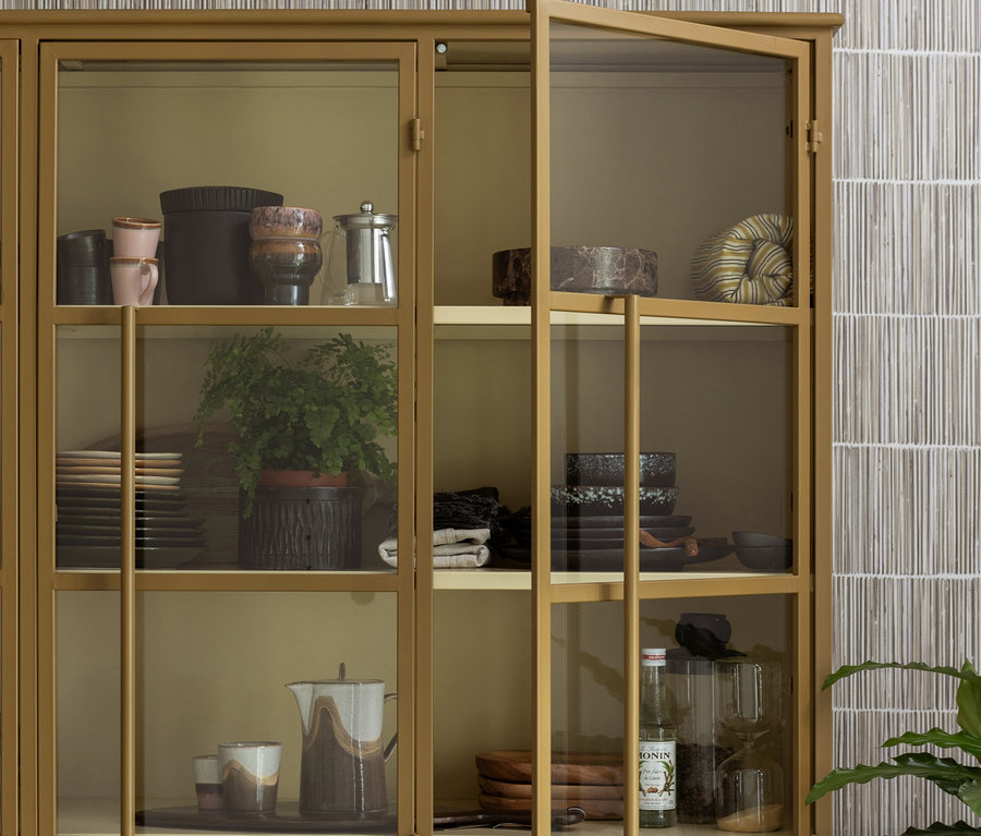Goldfarbene Vitrine mit Glastüren, eine davon offen, gefüllt mit Tassen, Schalen, einer Kaffeemaschine, einer Farnpflanze, Tellern, einem Keramikkrug und einer Sirupflasche.