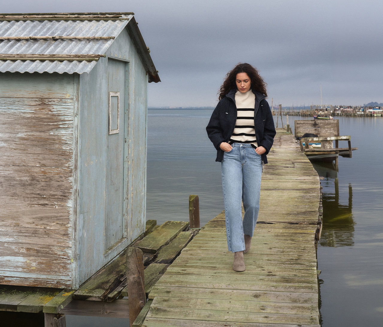 Frau geht auf einem Holzsteg neben einer verwitterten blauen Hütte, trägt einen gestreiften Pullover, eine dunkle Jacke und Jeans, mit ruhigem Wasser und bedecktem Himmel.