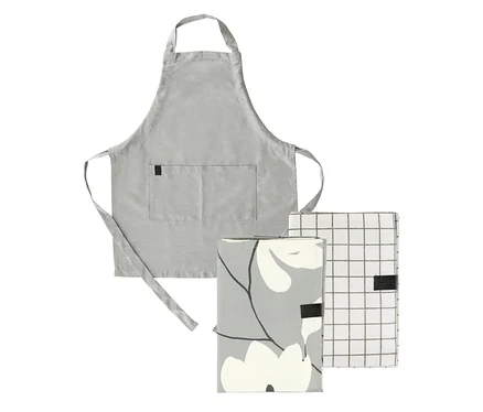 Eine hellgraue Schürze mit einer Vordertasche und Bändern, hinter zwei gefalteten Küchentextilien positioniert. Das linke Textil ist grau mit einem weißen Blumenmuster, das rechte ist weiß mit einem grauen Karomuster.