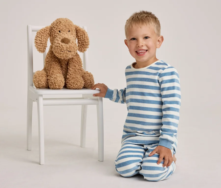 Ein lächelnder kleiner Junge in blau-weiß gestreiften Pyjamas kniet neben einem weißen Stuhl, auf dem ein brauner Plüschhund sitzt.
