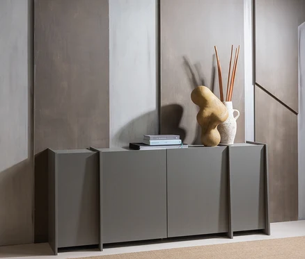Ein modernes graues Sideboard mit zwei Türen steht auf einem hellbraunen Teppich vor einer strukturierten grauen Wand. Darauf sind ein Stapel Bücher, eine abstrakte hellbraune Skulptur und eine weiße Vase mit Dekorationsstäben arrangiert.