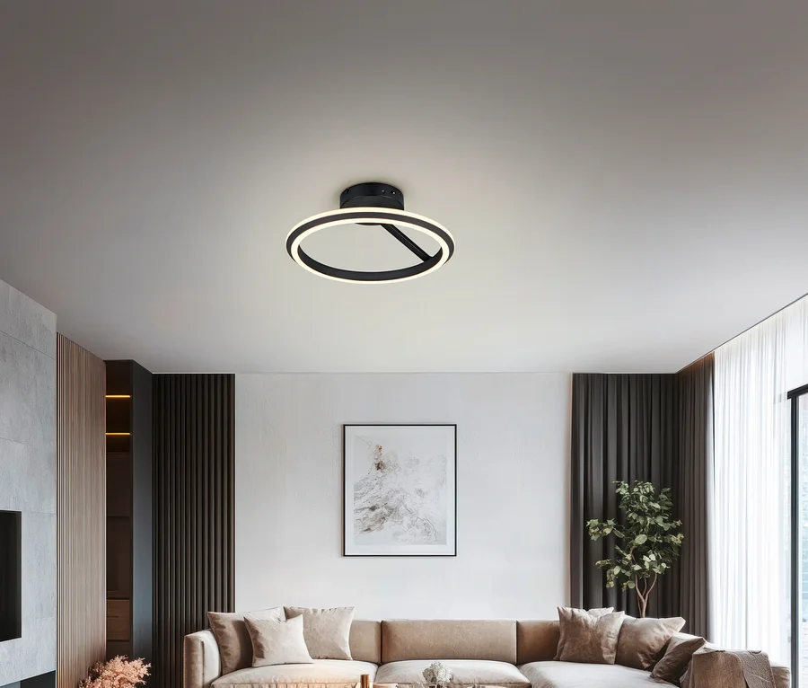 Modernes Wohnzimmer mit einer schwarzen, runden Deckenleuchte, die den Raum beleuchtet. Darunter steht ein großes beiges L-förmiges Sofa mit Kissen vor einer weißen Wand mit abstrakter Kunst. Dunkle Vorhänge rahmen ein Fenster rechts ein, daneben eine Topfpflanze.