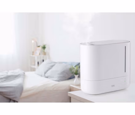 Weißer Luftbefeuchter, der Nebel auf einem Nachttisch in einem unscharfen Schlafzimmer abgibt.