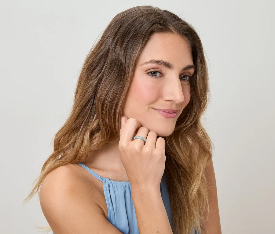 Frau mit gewelltem Haar und blauem Top berührt ihren Finger mit einem Ring vor einem hellen Hintergrund.