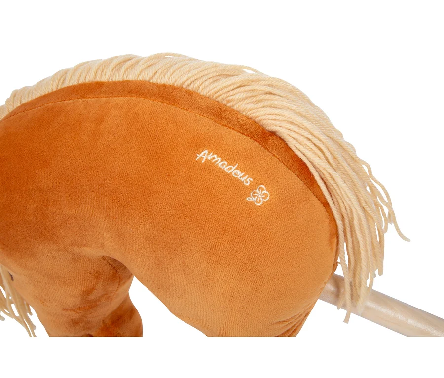 Nahaufnahme des Kopfes und Halses eines karamellfarbenen Plüsch-Steckenpferdes mit heller blonder Garnmähne. Der Name „Amadeus“ und ein kleines Blumensymbol sind weiß auf die Seite gestickt. Ein Holzstab ist darunter sichtbar.
