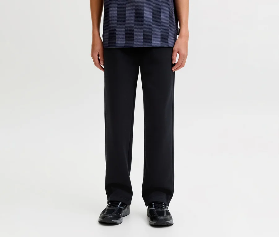 Eine Person steht und trägt eine schwarze Jack&Jones Bradley Sweatpants, ein dunkelblau gemustertes Shirt und dunkle Sneaker.