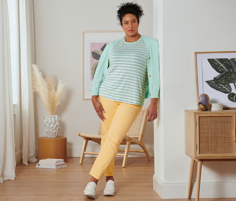 Eine Frau lehnt an einer Wand und trägt ein mintfarbenes, gestreiftes T-Shirt, einen Cardigan mit V-Ausschnitt, eine gelbe Slimfit-Jeans und Veloursleder-Sneaker.