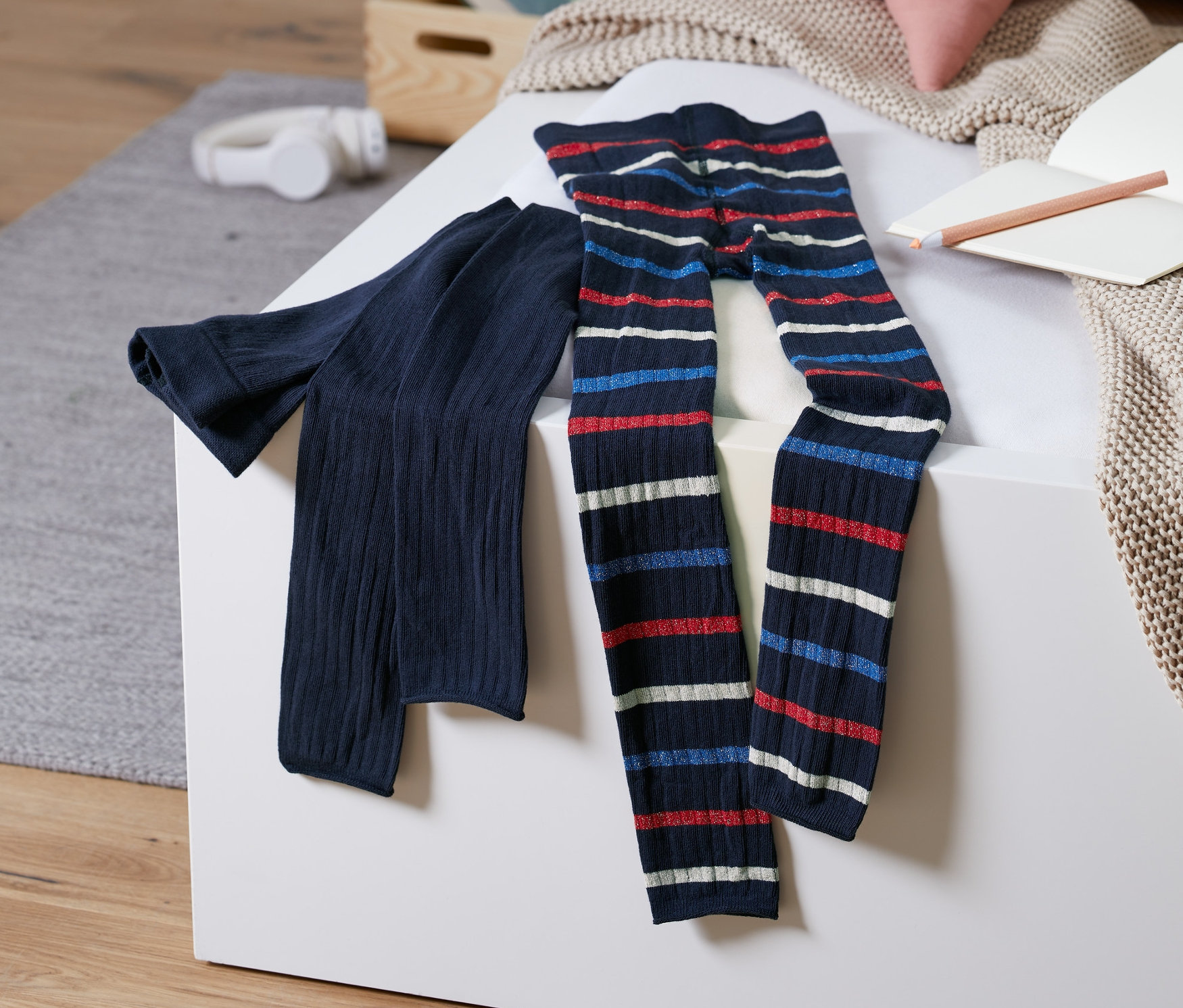 Zwei Strickleggings liegen auf einer weißen Oberfläche. Eine ist dunkelblau, die andere hat rot-weiß-blaue horizontale Streifen.
