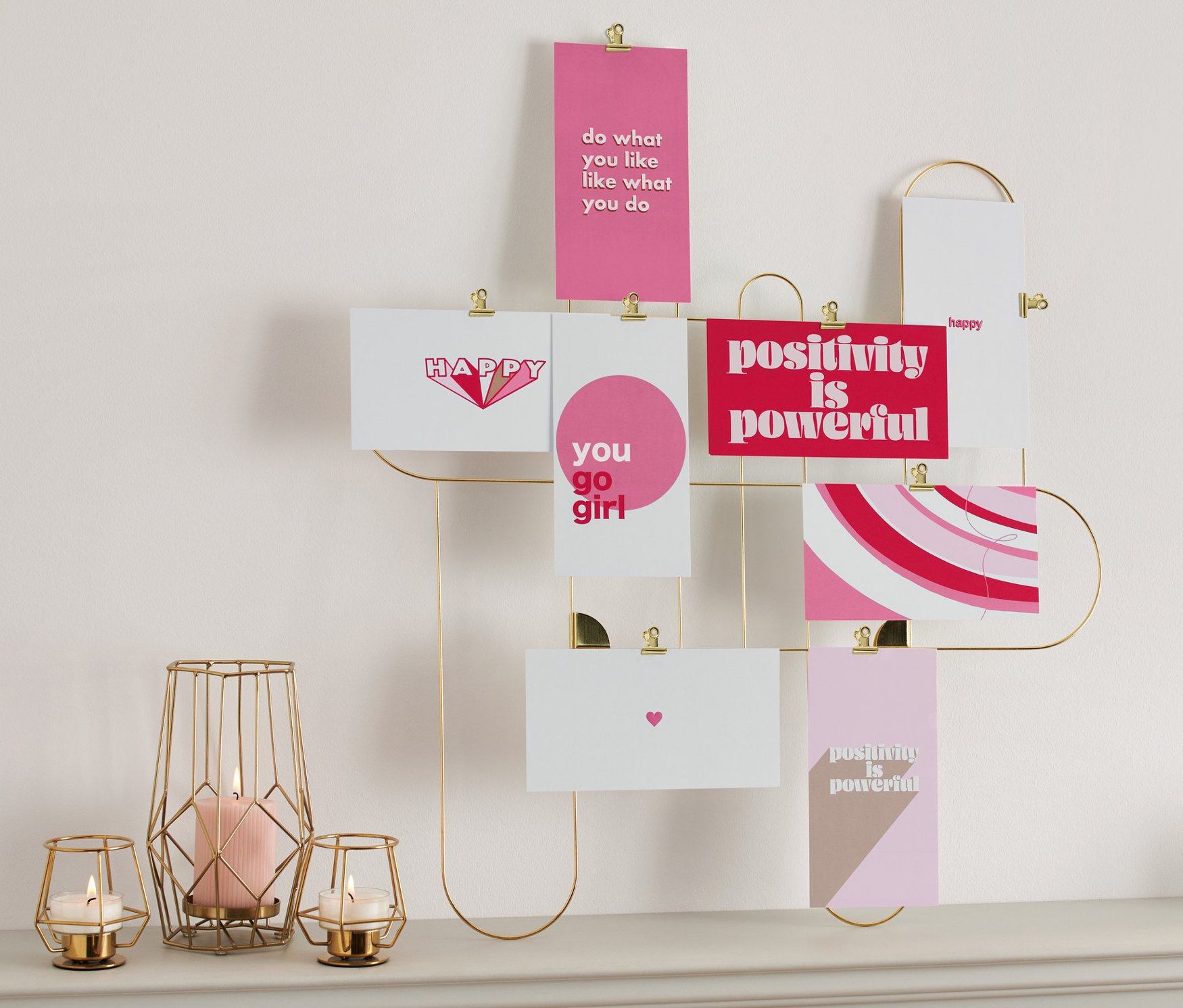 Postkarten-/Minibilder-Set hängen an einem goldenen Rahmen an der Wand über einer Tischplatte, auf der Kerzen und eine Uhr stehen.