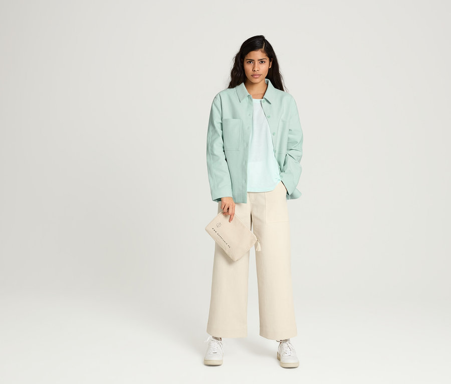Frau posiert in NAH/STUDIO Culotte aus ungefärbter Baumwolle, NAH/STUDIO Blusenjacke aus recycelter Baumwolle, NAH/STUDIO T-Shirt REFIBRA™ Ocean Blue und NAH/STUDIO Kosmetiktasche aus Baumwolle.
