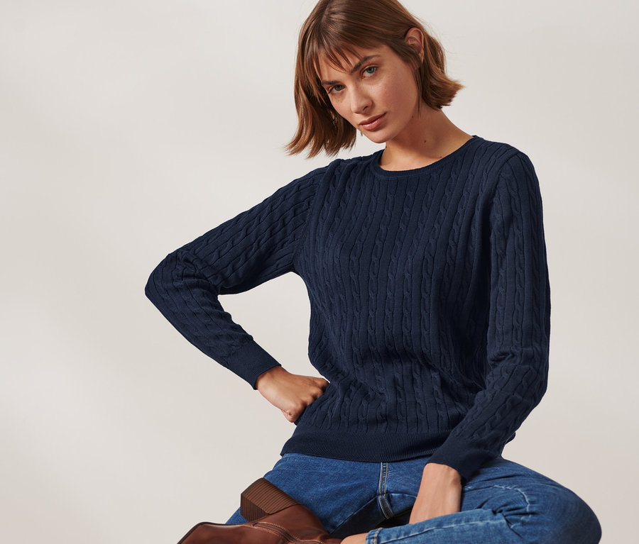 Frau sitzt auf einem Hocker und trägt einen dunkelblauen Zopfstrickpullover.
