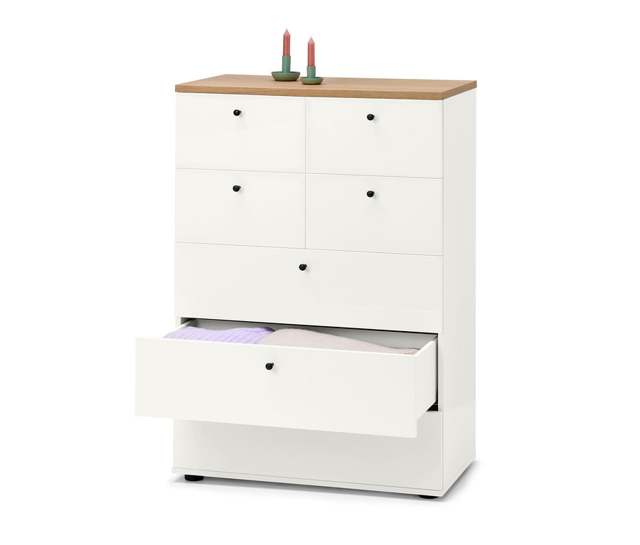 Eine weiße Schubladenkommode "Bekeson" mit sechs Schubladen, von denen eine offen ist und violette und beige Pullover zeigt. Auf der Kommode stehen zwei grüne Kerzen.
