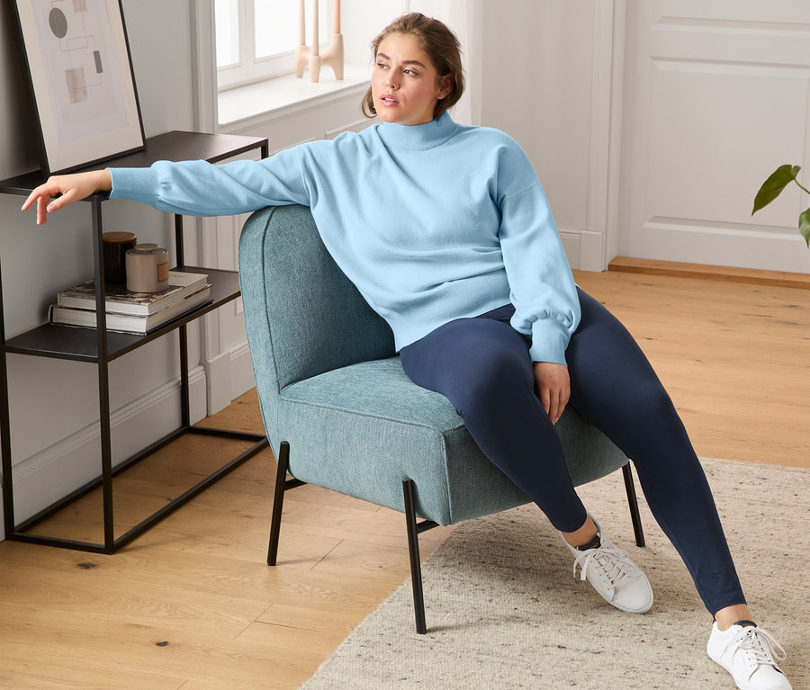 Eine Frau sitzt in einem Sessel und trägt Sneaker aus chromfrei gegerbtem Leder, Leggings und einen Pullover mit Ballonärmeln.