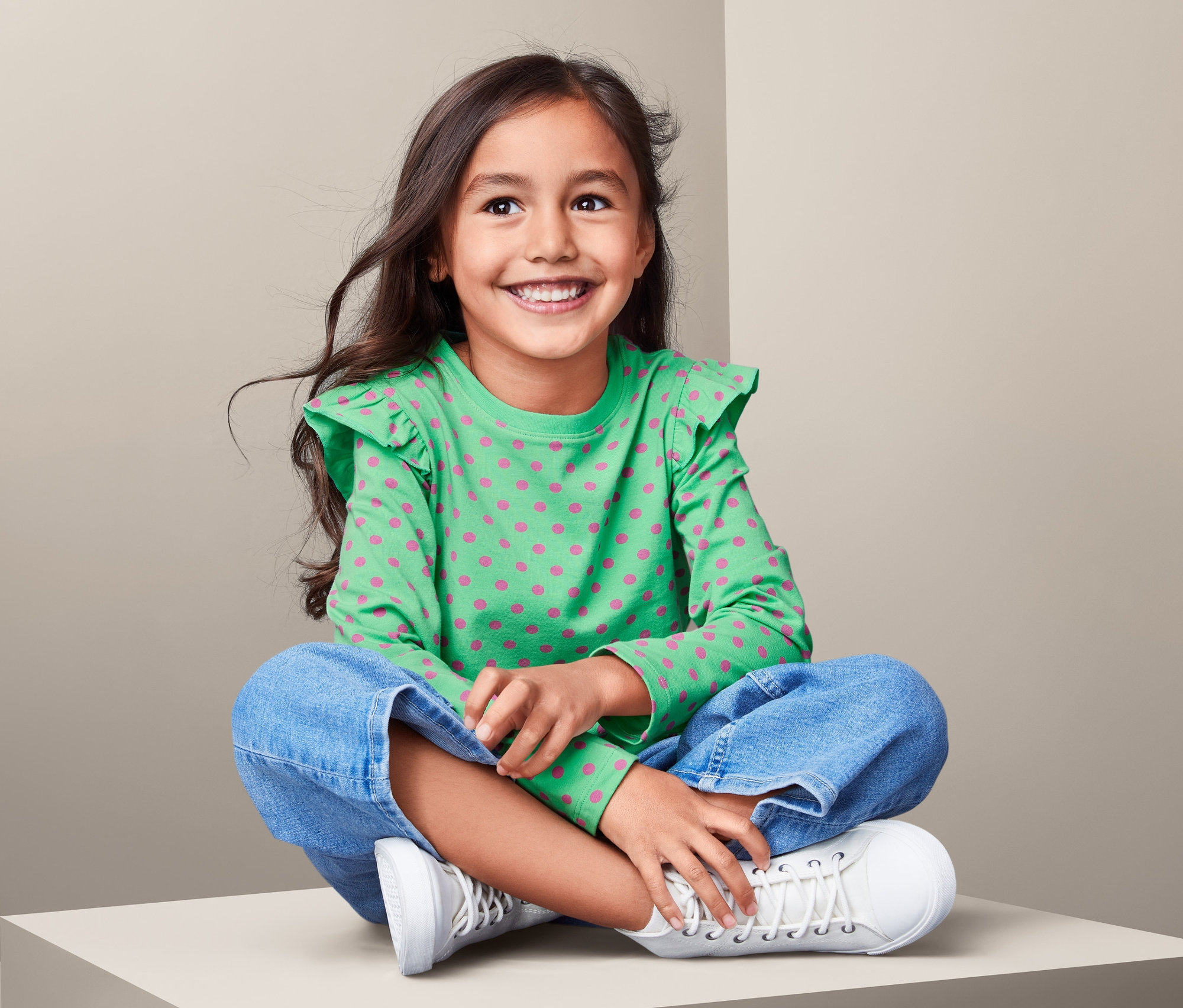 Ein lächelndes Mädchen sitzt mit gekreuzten Beinen auf einem Sockel und trägt ein grünes Langarmshirt mit rosa Punktmuster.
