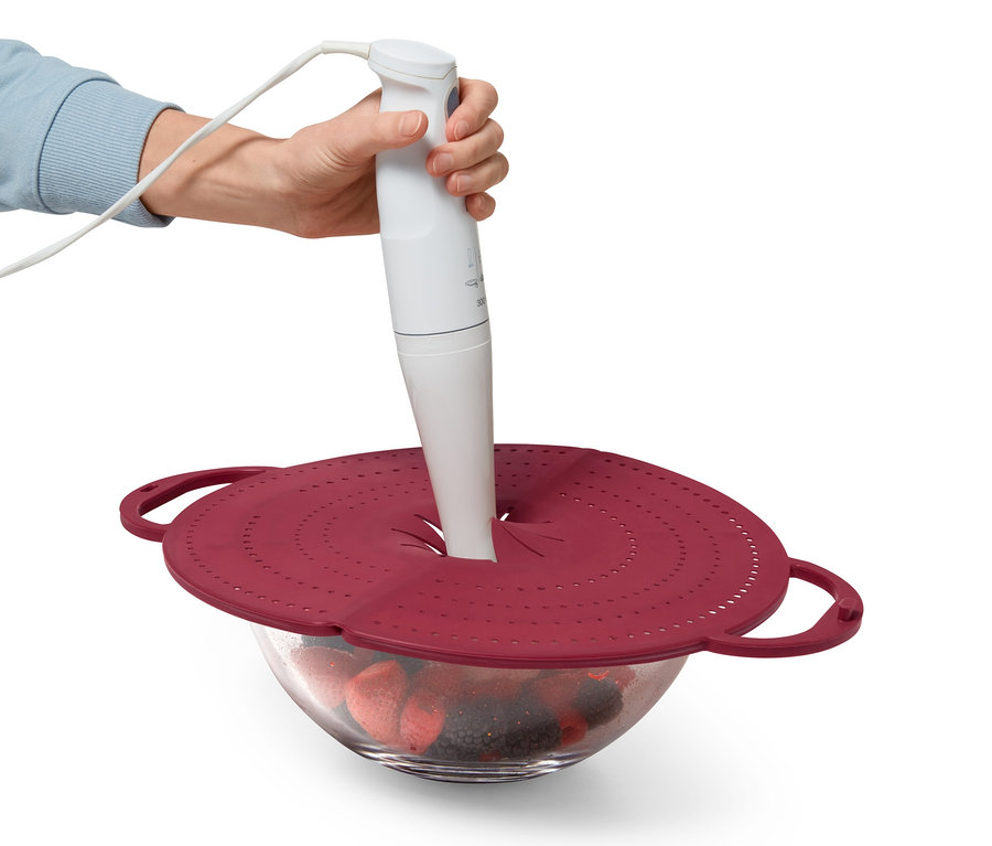 Eine Hand hält einen Stabmixer über eine Schüssel mit Früchten, die mit einem roten 4-in-1-Pfannenspritzschutz abgedeckt ist.