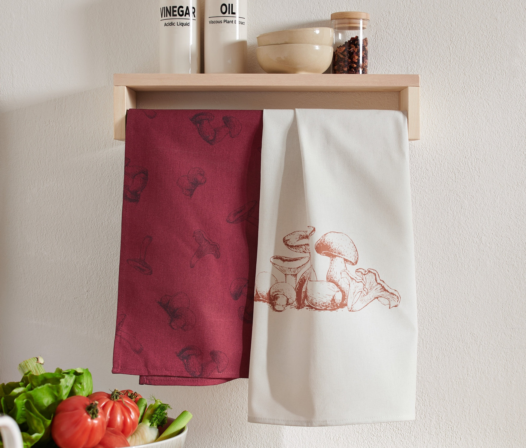 Zwei Geschirrtücher hängen an einem Holzregal. Eines ist weinrot mit einem Pilzaufdruck, das andere ist weiß mit einer Pilzillustration.
