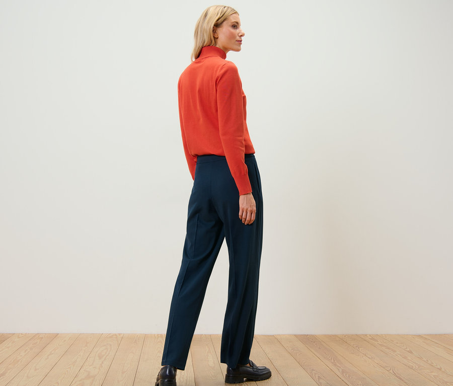 Eine Frau steht mit dem Rücken zur Kamera und trägt einen orangefarbenen Rollkragenpullover und eine marineblaue Jerseyhose mit Biese.