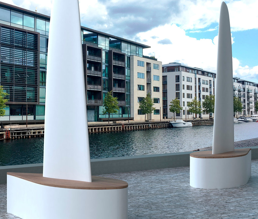 Zwei weiße Wings for Living Bank »Sydney« Bänke in Segelbootform stehen an einer Uferpromenade mit Gebäuden und Wasser im Hintergrund.