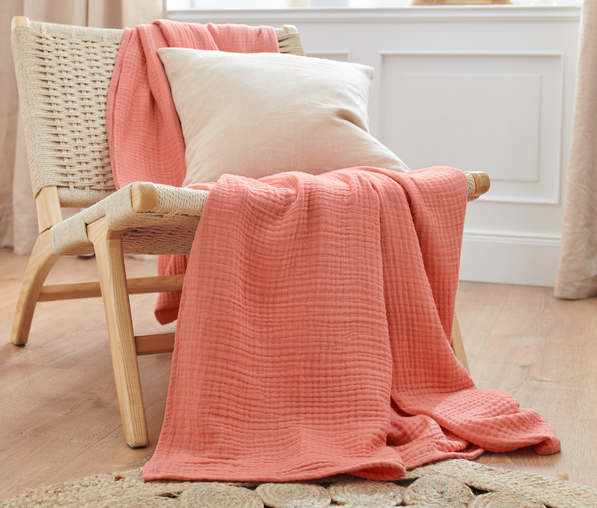 Eine pfirsichfarbene Tagesdecke und ein cremefarbenes Kissen liegen auf einem Korbstuhl.