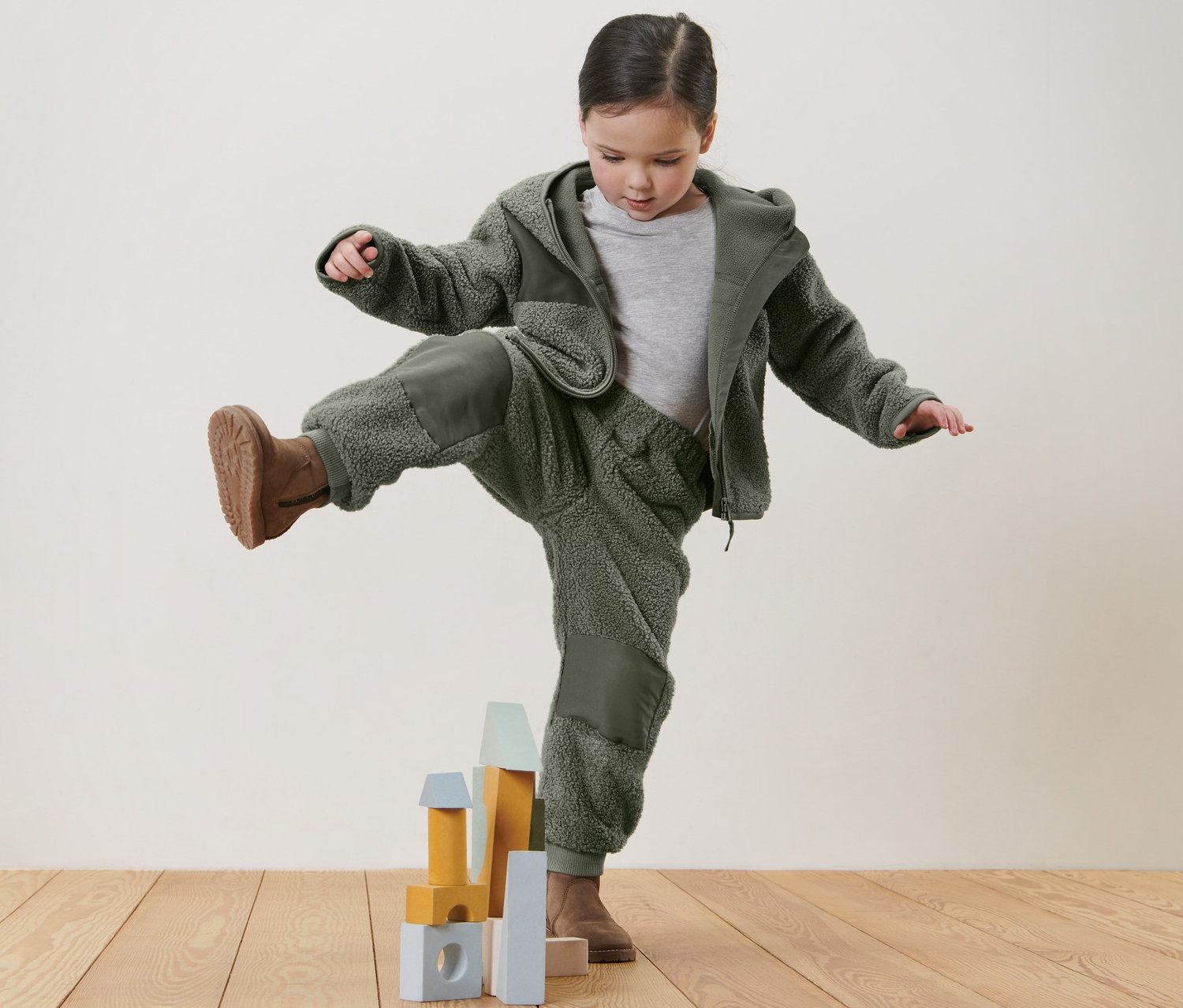 Mädchen steht auf einem Bein auf einem Holzboden und spielt mit Kinder-Teddy-Hose und Bauklötzen.