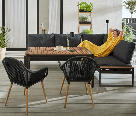 Eine Frau im gelben Kleid liegt auf der Dining-Lounge-Ecke »Elin« mit Premium-Komfort-Kissen. Im Vordergrund stehen zwei Schalenstühle mit Textilgeflecht und ein Tisch. Im Hintergrund steht ein Premium-Grillwagen »Elin«.