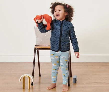 Lächelndes kleines Mädchen hält Plüschfuchs. Sie trägt blaue Leggings mit Wolken und eine blaue Sweatshirtjacke.