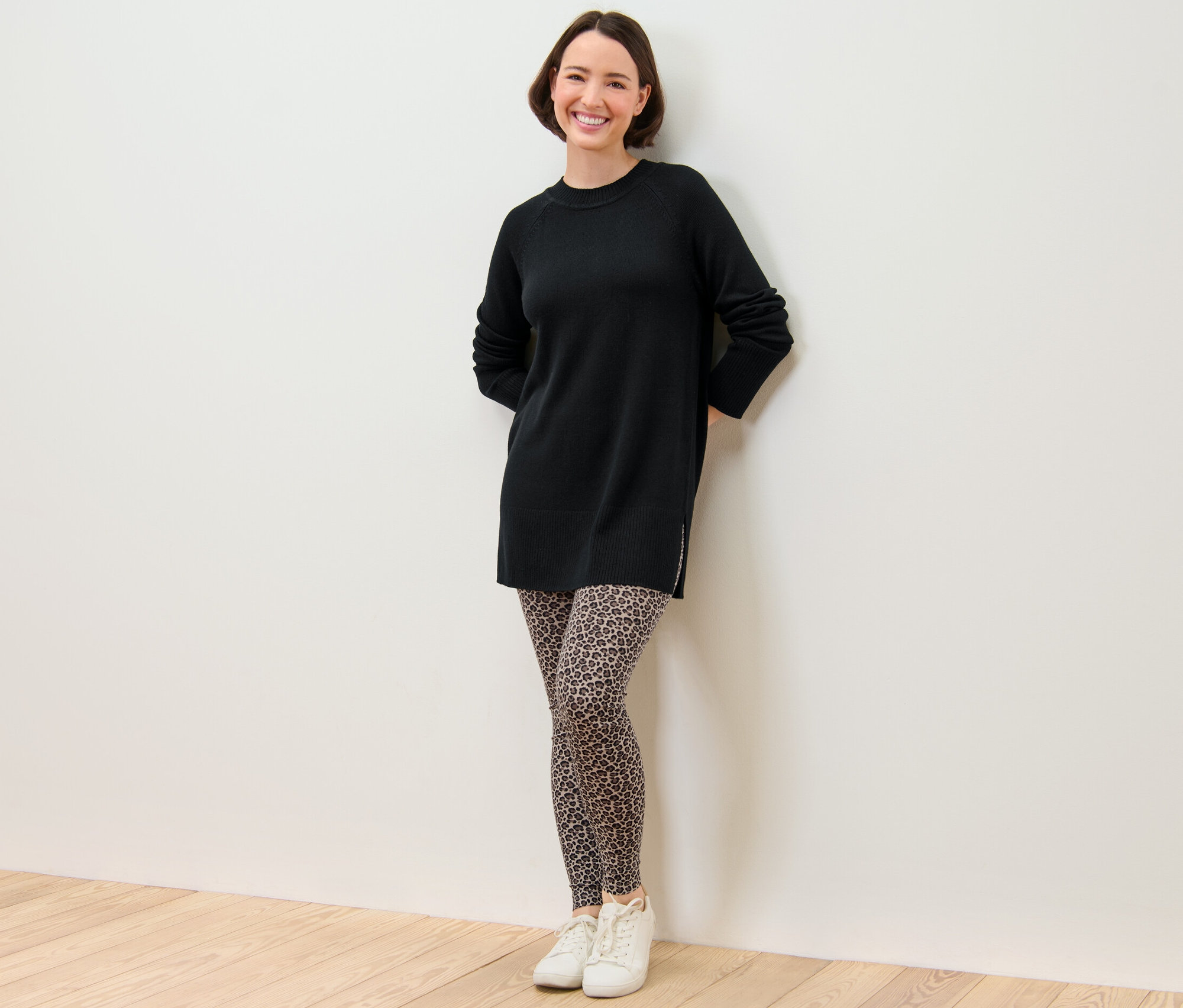 Eine Frau steht und lächelt, sie trägt ein schwarzes Kleid und bedruckte Leggings.