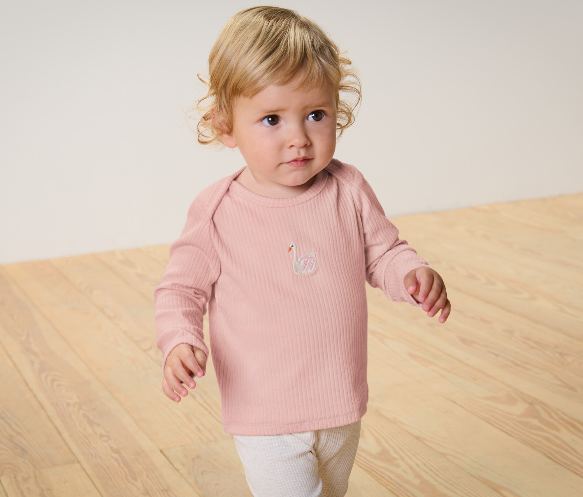 Ein Baby steht auf einem Holzboden und trägt ein rosa Langarmshirt mit Rippstruktur und eine weiße Hose.