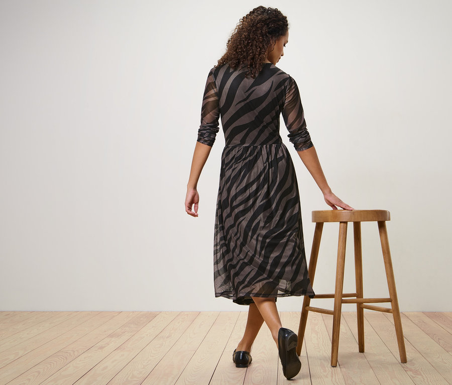 Frau posiert in bedrucktem Mesh-Kleid und stützt sich mit einer Hand auf einen Holzhocker.