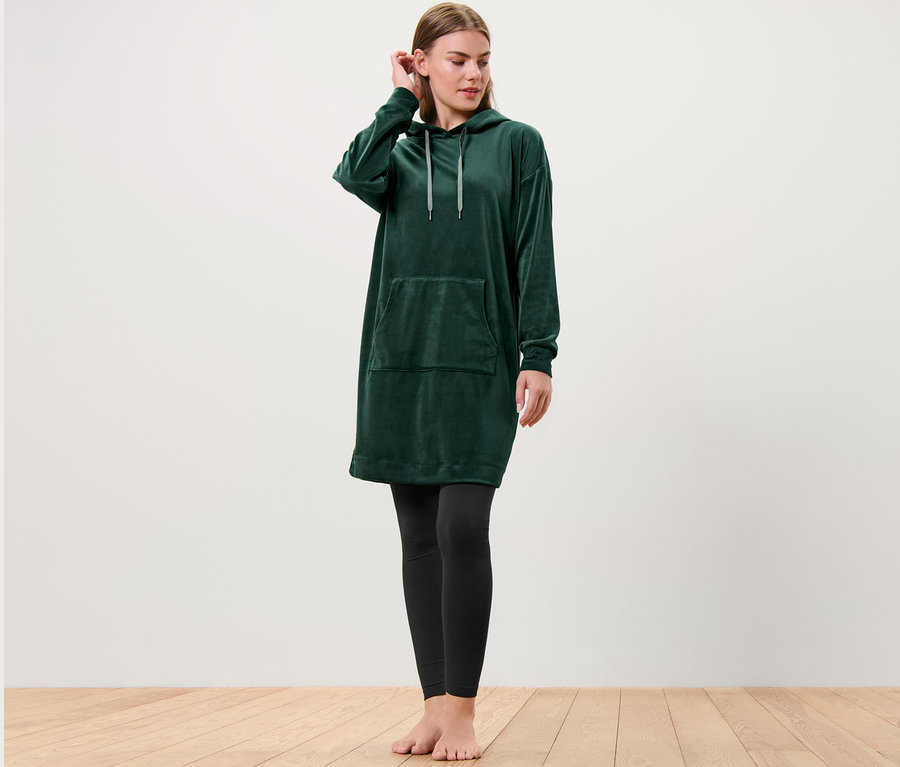 Frau steht barfuß und trägt ein Nicki-Loungewear-Kleid in Grün mit Kapuze und eine Leggings in Schwarz.