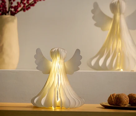 Zwei kleine LED-Papier-Engel, einer auf einem Schrank und der andere auf einem Regal, neben einer Vase mit roten Beeren und einem Teller mit Nüssen.
