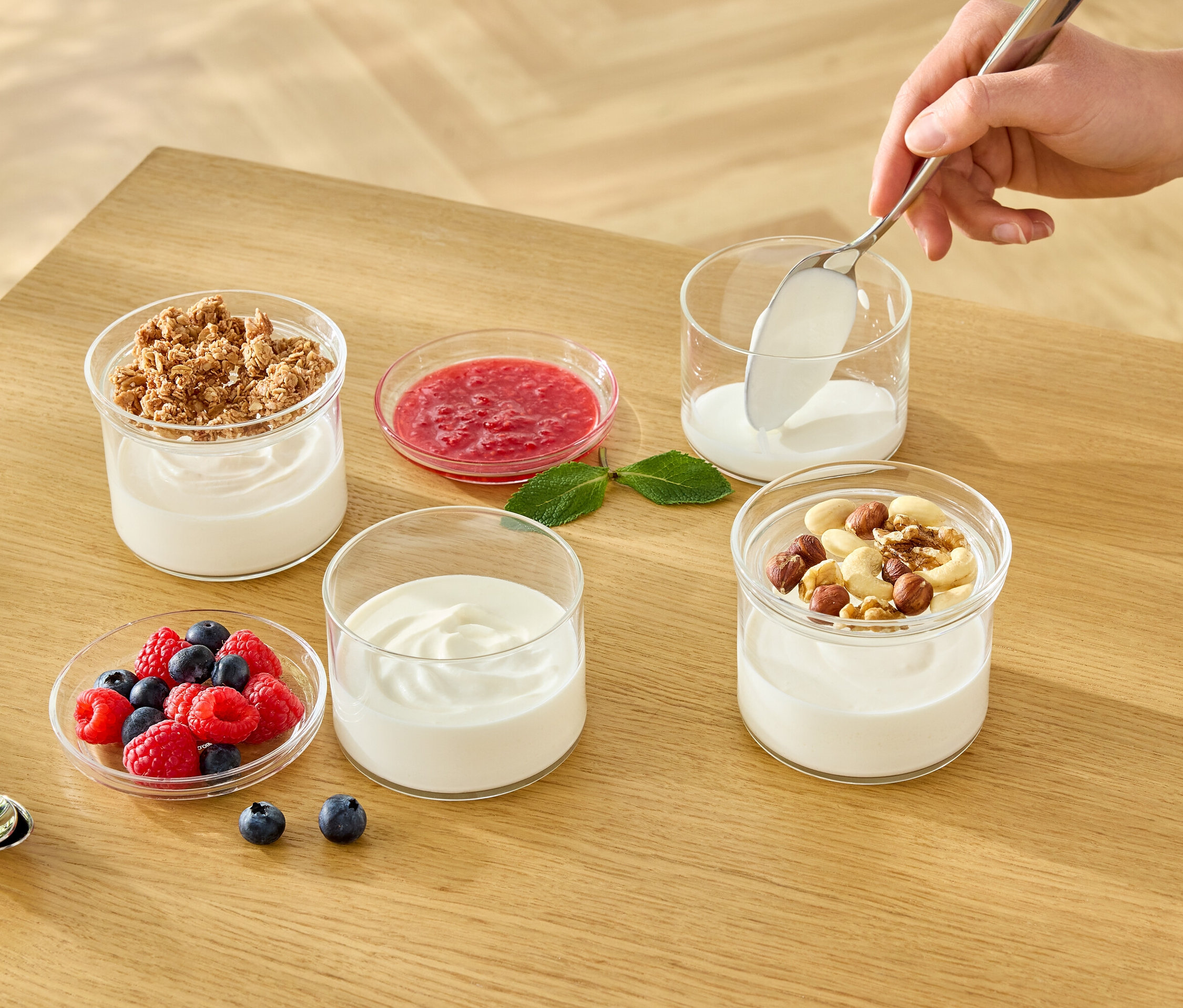 Eine Hand löffelt weißen Joghurt in ein Glas. Auf dem Tisch stehen weitere Gläser mit Joghurt, Müsli, Früchten und Nüssen.
