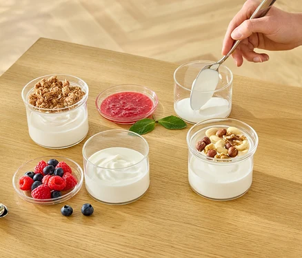 Eine Hand löffelt weißen Joghurt in ein Glas. Auf dem Tisch stehen weitere Gläser mit Joghurt, Müsli, Früchten und Nüssen.