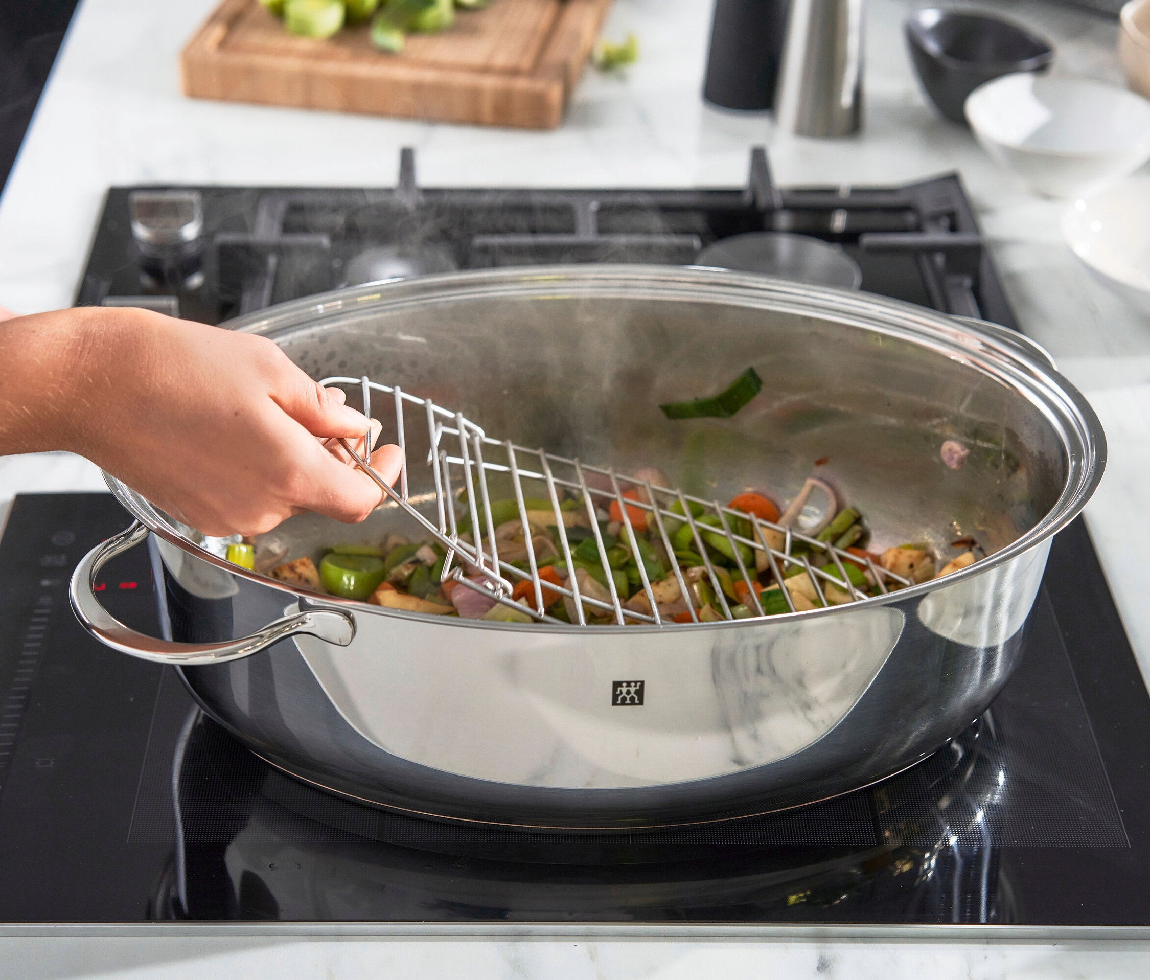 Eine Hand hält ein Edelstahlgitter über eine Pfanne mit sautiertem Gemüse auf einem Induktionskochfeld.