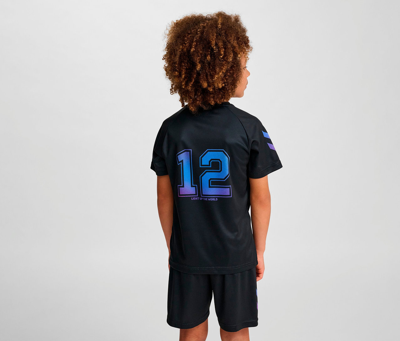 Junge mit lockigem Haar trägt ein schwarzes HUMMEL HMLJR shimmer T-Shirt S/S und schwarze Shorts, steht mit dem Rücken zur Kamera.
