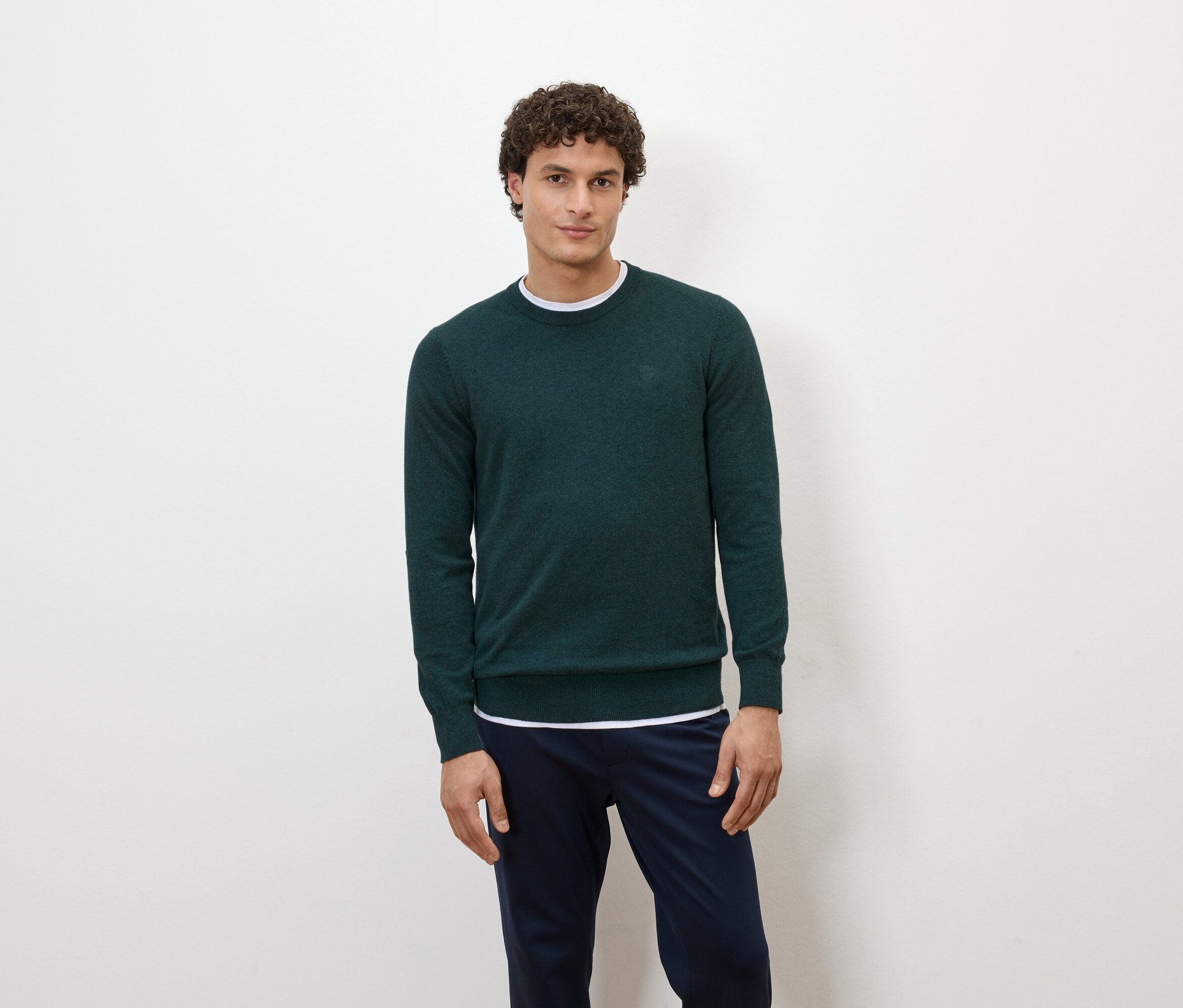 Ein Mann posiert vor einem weißen Hintergrund in einem dunkelgrünen Pullover und dunkelblauen Hosen.
