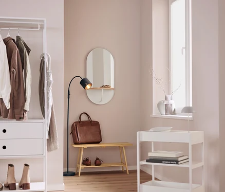 In einem Interieur befinden sich ein weißes Regal mit Kleidung, ein ovaler Spiegel, eine braune Ledertasche auf einer Bank, eine schwarze Lampe und ein weißer Rollwagen.