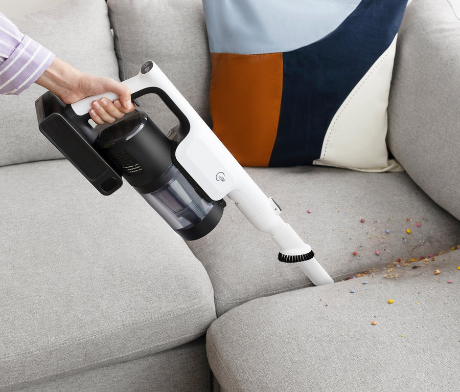 Eine Hand hält einen Tineco Akku-Staubsauger »Pure One A50S« und saugt Krümel von einem grauen Sofa.