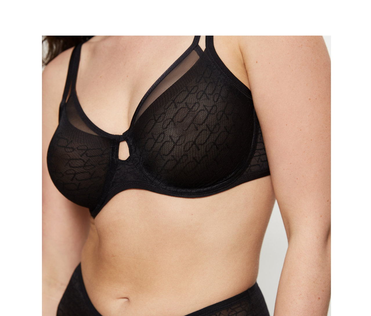 Frau trägt einen schwarzen Triumph Signature Sheer Minimizer BH und passenden, gemusterten Slip. Der BH hat einen kleinen Ausschnitt zwischen den Cups.